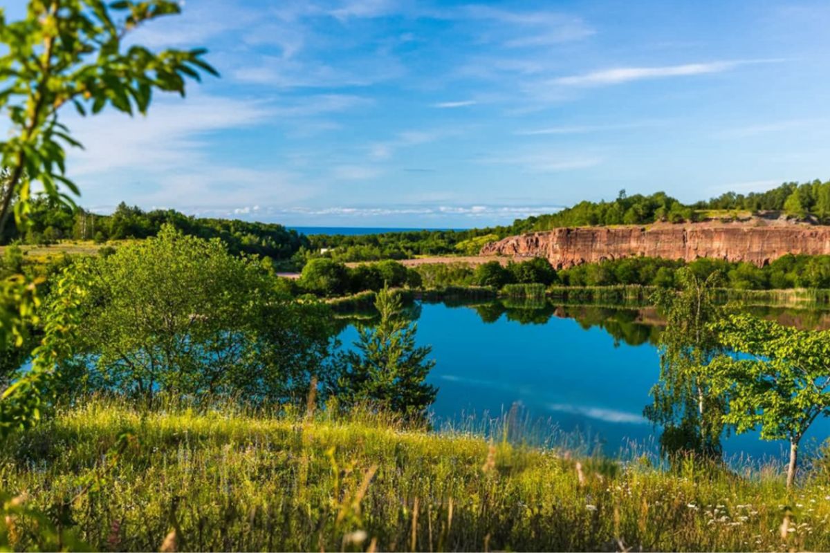 Sjö och grönskande landskap med Kinnekulles röda klippor i bakgrunden under en klarblå himmel.