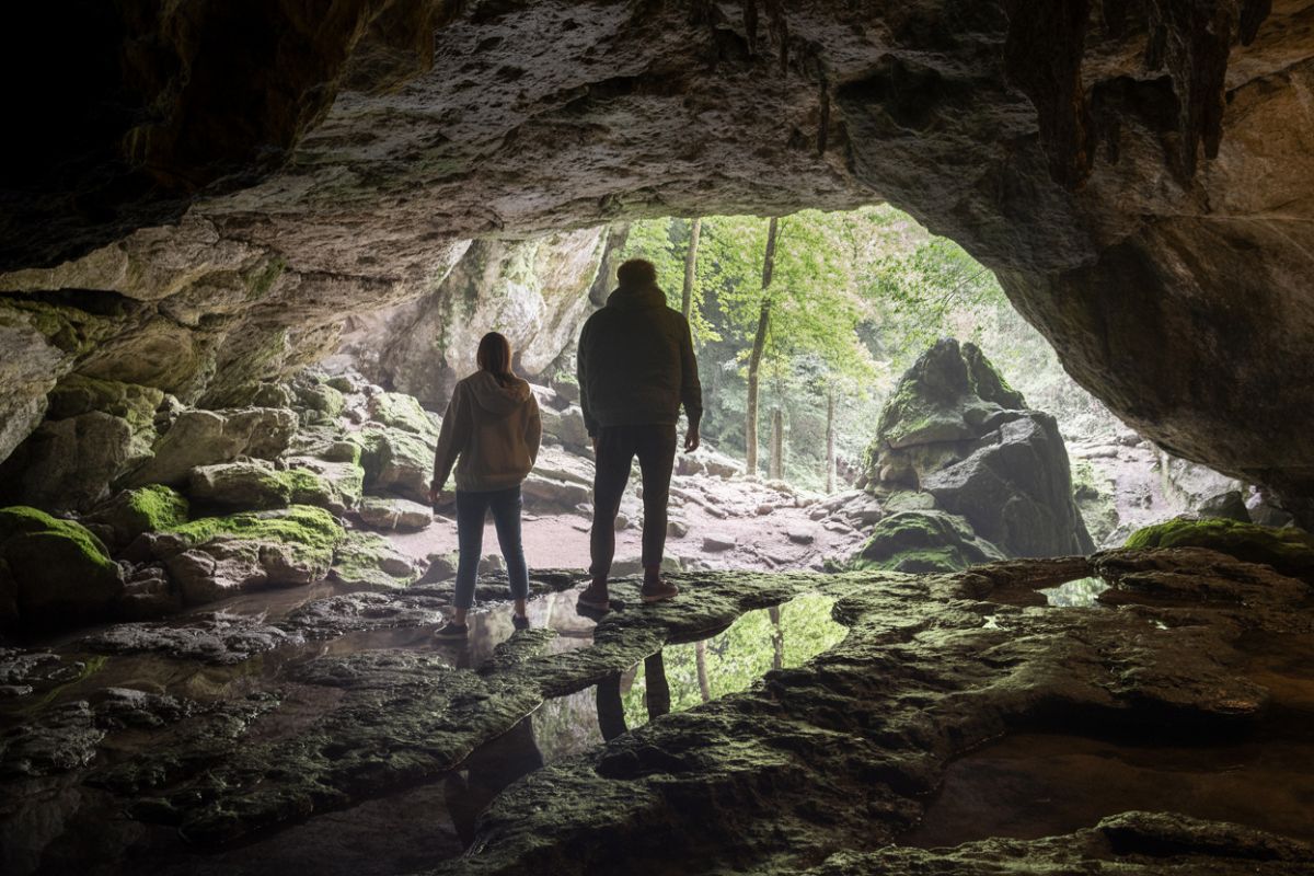Grottor att uppleva vid vandring Vildmarknaden.se Två personer står vid öppningen av en grotta med utsikt mot grönskande skog, grottväggar och mossbeklädda stenar.