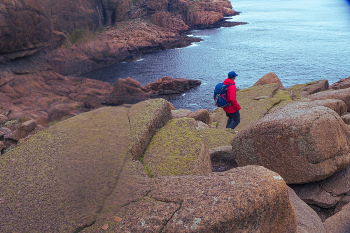 Vandra vid havet i Sverige Vildmarknaden.se Person med röd jacka och ryggsäck vandrar över stora klippor vid havet.